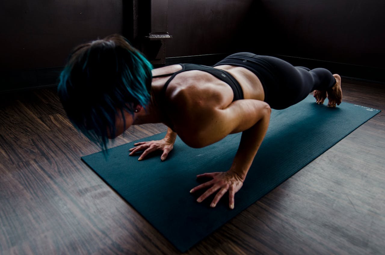 gallery-1 A fit woman with blue hair performs a push-up on a yoga mat indoors, showcasing strength and focus.