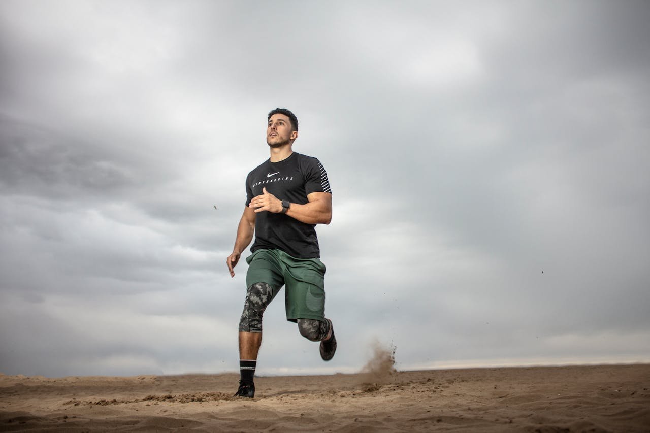 gallery-4 A muscular man running on sandy terrain under cloudy skies, promoting fitness and adventure.